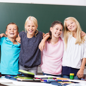 Eine Gruppe lächelnder Mädchen vor eine Schultafel. Im Vordergrund ein Schreibtisch mit Federmappe und Stiften. Link zur Rubrik Bildung